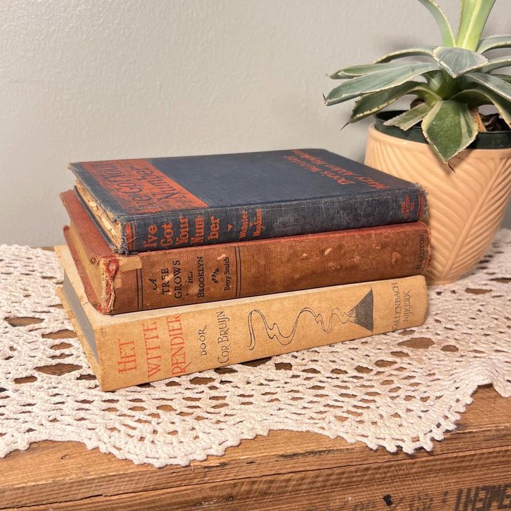 A stack of three old books on top of w doily on a wooden table, beside a potted plant.