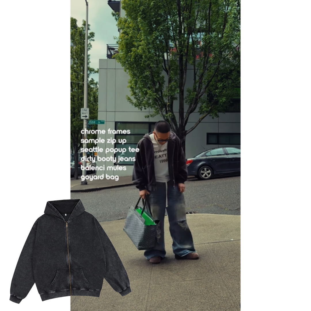 Image of Duc Le posing in a the street. He is wearing a washed black zip up hoodie, a white tshirt, and sneakers. There is a building and large green tree in the background.