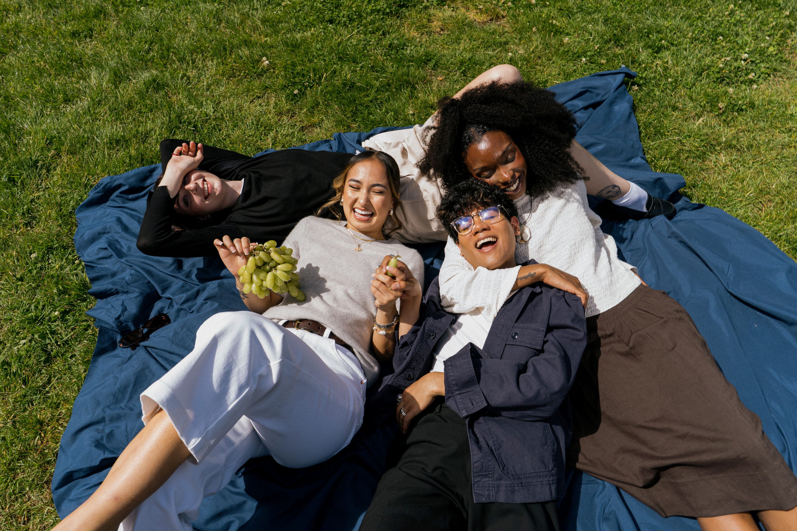 Four friends laying down on a picnic mat laughing and hanging out wearing summer clothes.