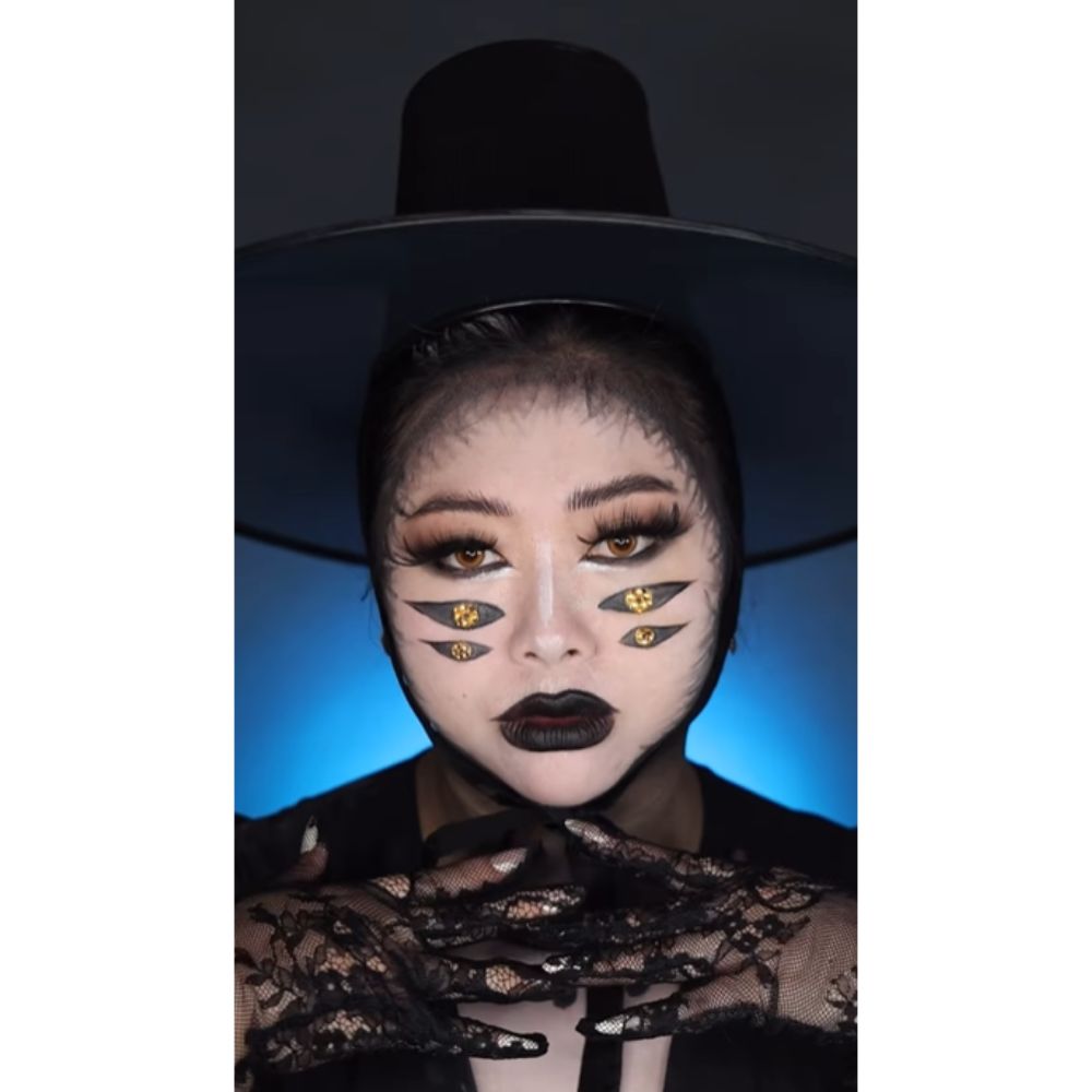 Influencer Lexy Bach dressed as the three eyed raven from the K-pop Demon Hunter movie. She has two extra sets of eyes painted below her actual eyes, black lipstick, a black top hat, black top, and lacy black gloves.