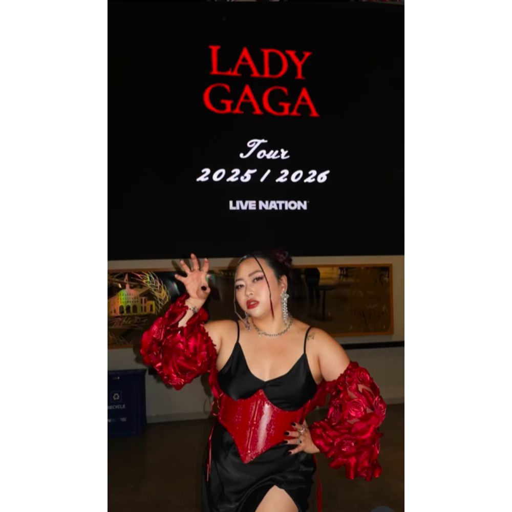 Influencer Lexy Bach posed in front of a sign that says "Lady Gaga Tour 2025\2026 Live Nation". Lexy is wearing a black silk dress, red corset, and red puffy sleeves.