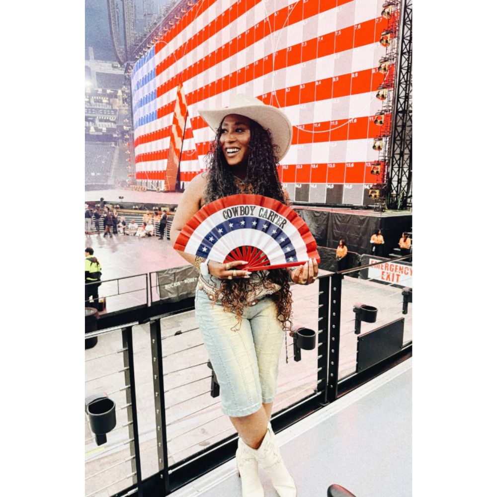Tash Haynes posting in front of an American Flag billboard at a Beyonce concert. She is wearing a white cowboy hat, light wash jorts, and white cowboy boots. She is holding an red, white, and blue fan that reads "COWBOY CARTER"