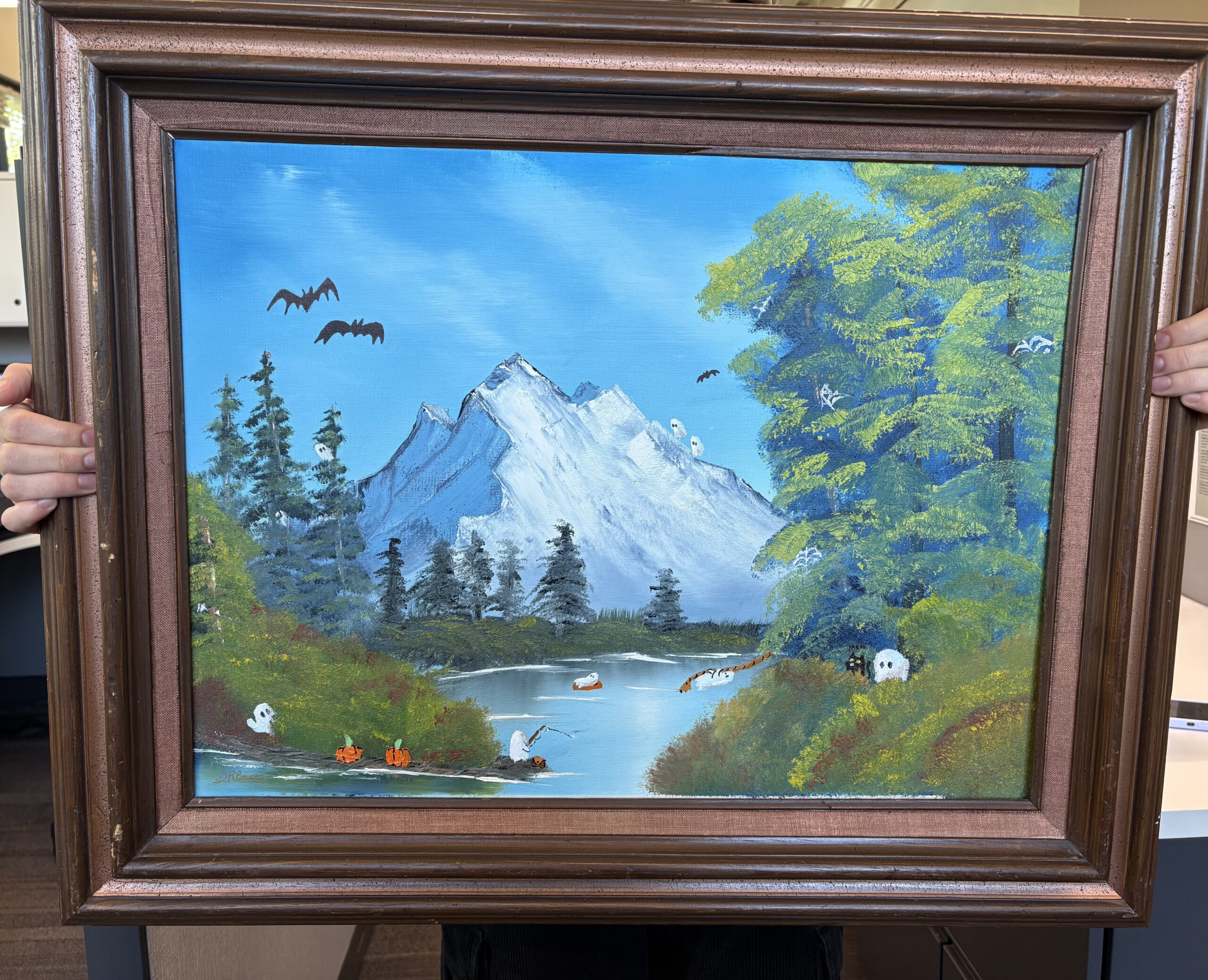 A large painting in a rectangular wooden frame. The painting is of a landscape of a mountain by a lake and trees. There are extra small bats, ghosts, and pumpkins painted onto the original painting.