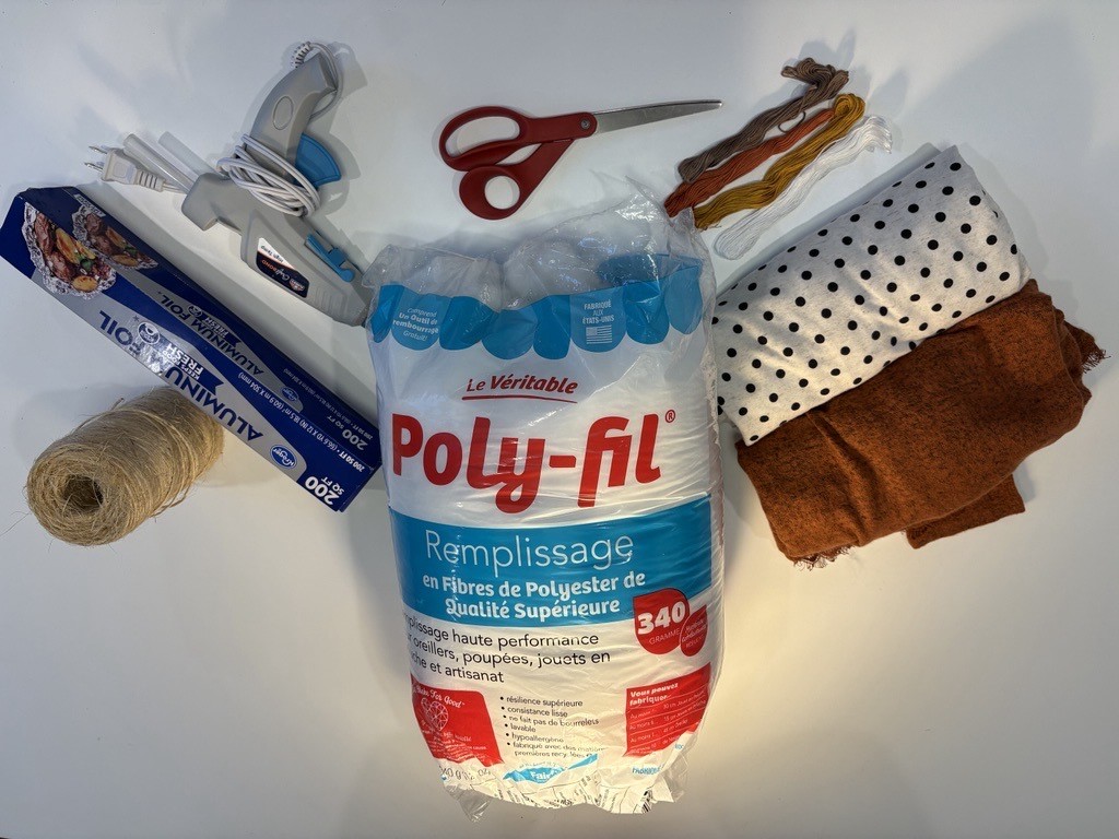 A hot glue gun, roll of twine, yarn, scissors, aluminum foil, Poly-fil, a rolled up polka dot tshirt, and an orange rolled up tshirt displayed on a white table.