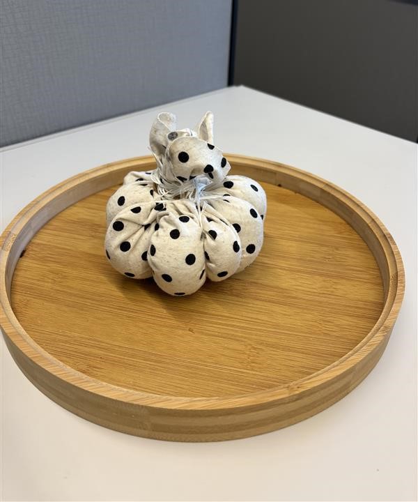 A black and white polka dot ball tied at the top with a rubber band. There yarn wrapped vertically around the ball several times in different directions to create ridges on the ball resembling a pumpkin. The ball is sitting on a circular brown wooden tray, which is placed on top of a white table.