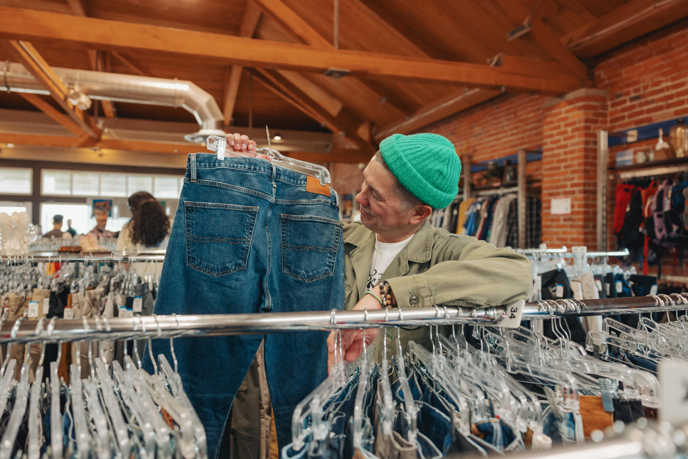 Brian Rauschenbach standing inbetween men's jeans aisle's in a Goodwill store. He is holding up and looking at a pair of denim jeans.