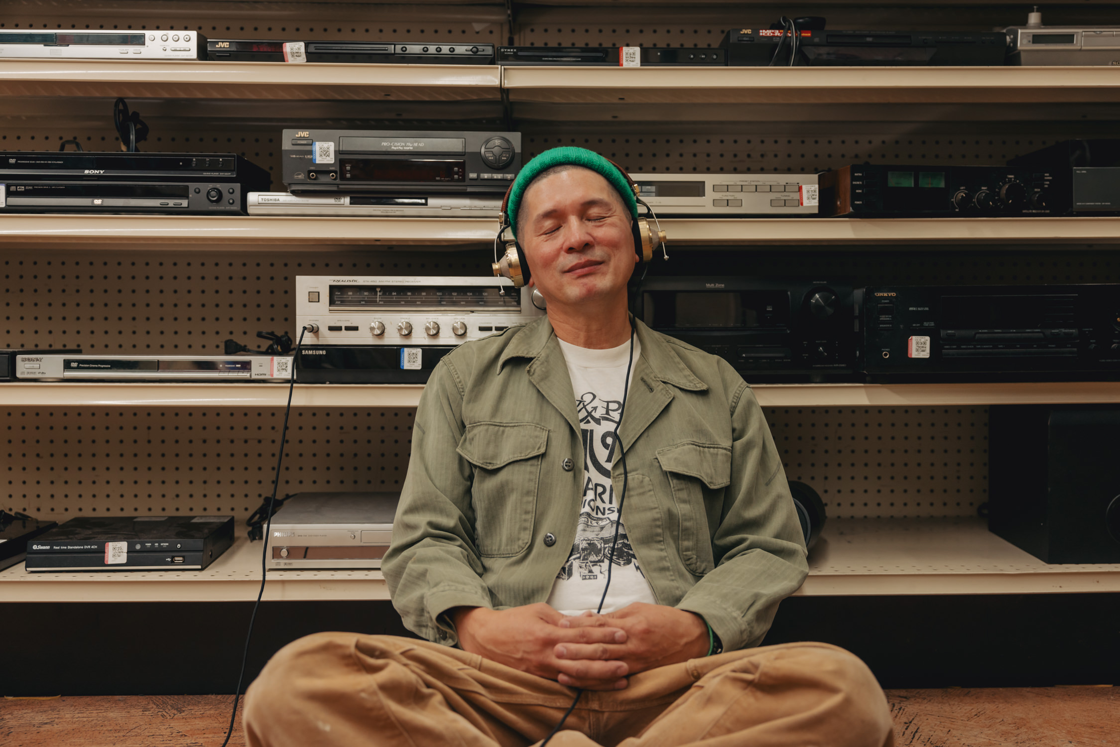 Brian Rauschenbach sitting in front of an aisle of audio equipment in a Goodwill store. His eyes are shut and he is wearing vintage looking headphones.