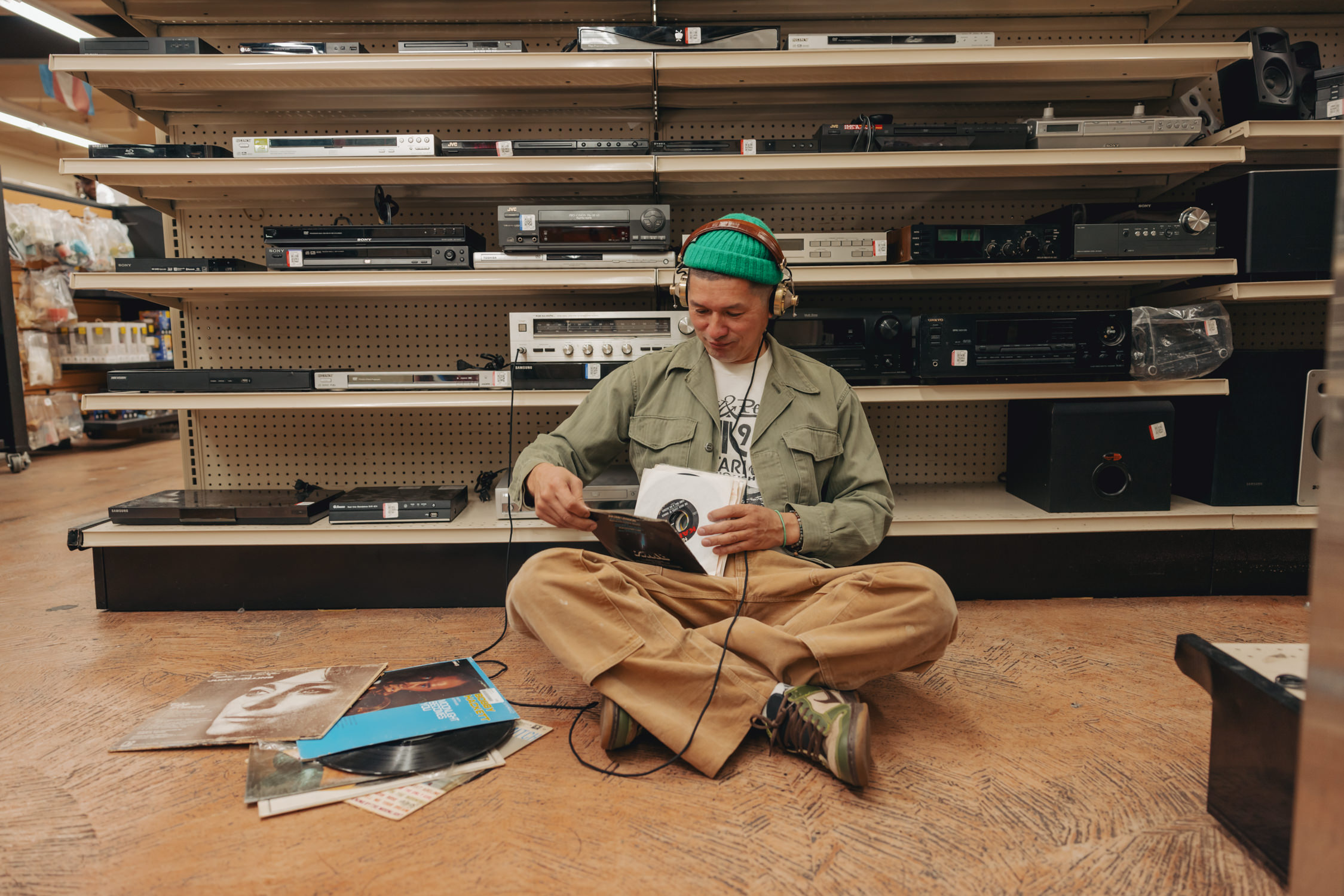 Brian Rauschenbach sitting on the floor of a Goodwill store in front of a wall of audio equipment. He is wearing vintage looking headphones and is actively sifting through vinyls.